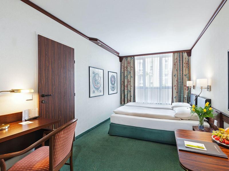 Světý hotelový pokoj se zeleným kobercem, manželskou postelí, psacím stolem a velkým oknem se závěsy.