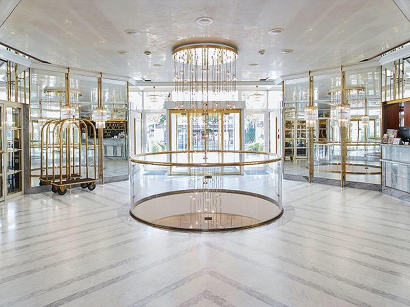 Luxusní hotelová hala s lustrem, skleněnými stěnami a moderní bílou mramorovou podlahou.