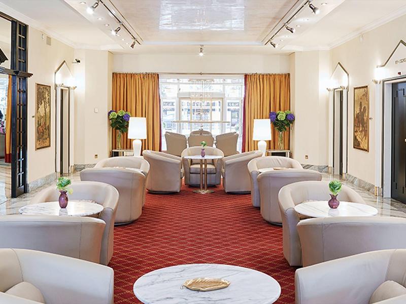 Jasný hotelový salonek s bílými křesly, červeným kobercem a velkými okny s oranžovými závěsy.