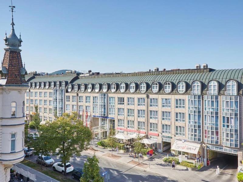 Elegantní hotelová budova s prosklenou střechou u slunné ulice se stromy.