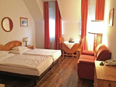 Acogedora habitación de hotel con dos camas individuales, sofá, mesa y cortinas naranjas.