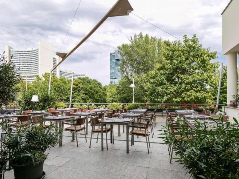 Terraza moderna al aire libre de un hotel con mesas, sillas y entorno verde bajo cielo nublado.