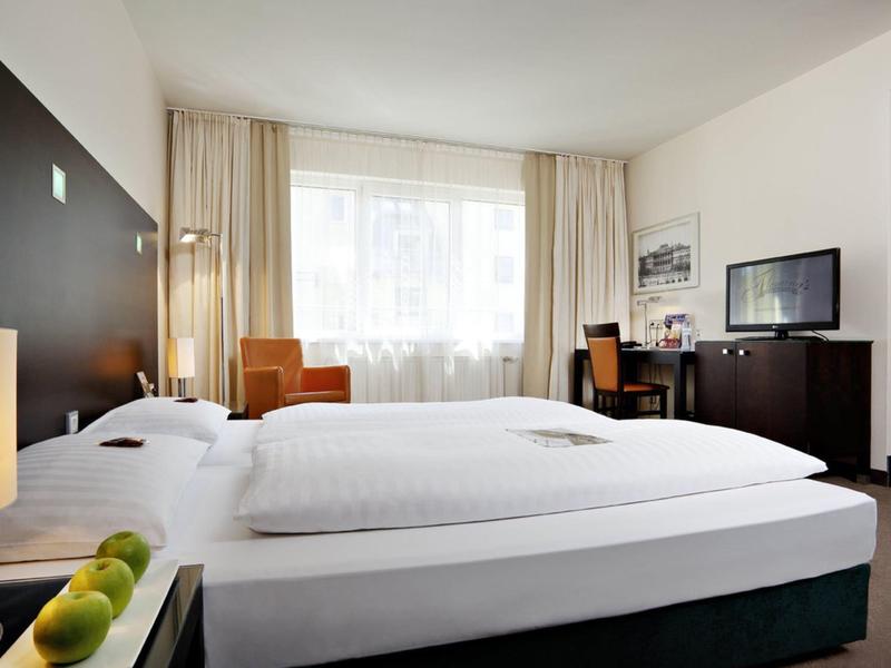 Chambre d'hôtel lumineuse avec deux lits simples, grande fenêtre, bureau et télévision sur meuble sombre.