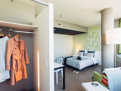 Habitación de hotel moderna con armario abierto y cama doble, colores claros y luz natural.