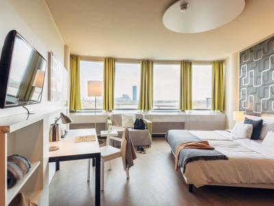 Hell eingerichtetes Hotelzimmer mit zwei Betten, Schreibtisch, Fernseher und großen Fenstern mit Vorhängen.