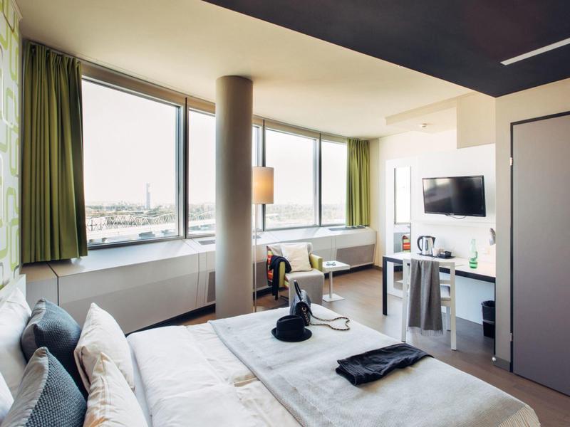 Habitación de hotel moderna con ventana grande, cama, televisor y cortinas verdes.