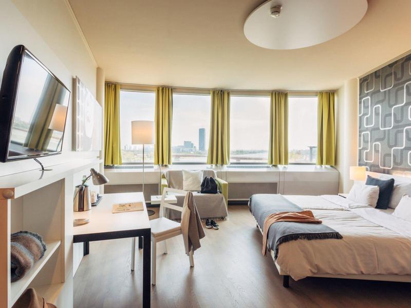 Habitación de hotel luminosa con dos camas, escritorio y grandes ventanas con vista a la ciudad.