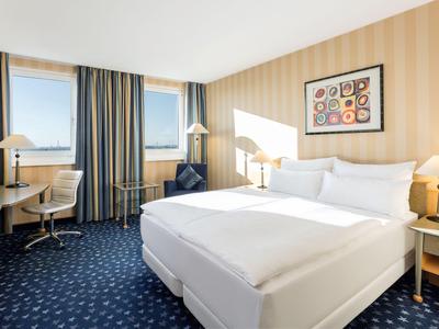 Světlý hotelový pokoj s manželskou postelí, pracovním stolem a okny se závěsy.