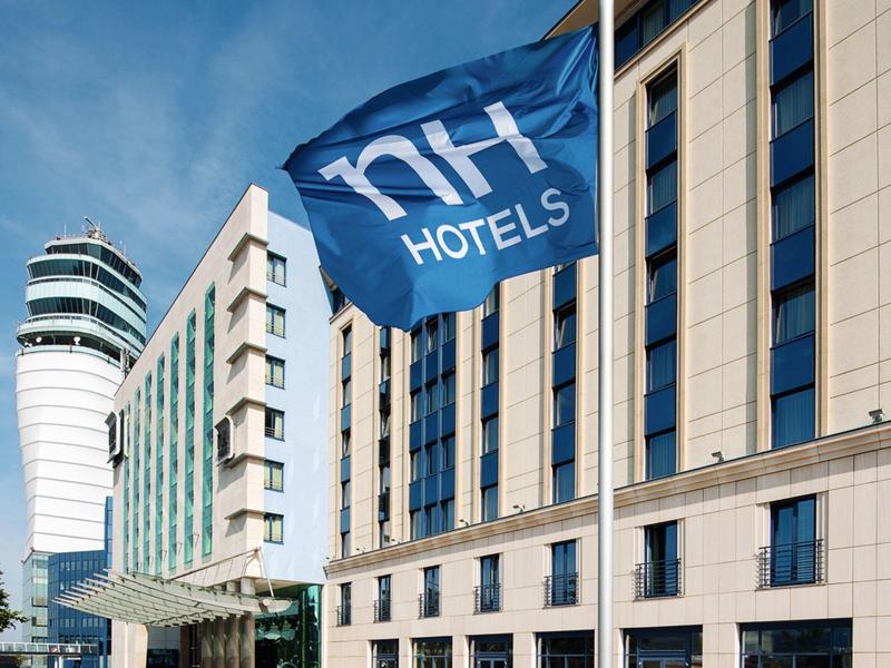 Außenansicht eines modernen NH Hotels mit blauer Flagge und mehreren weißen Autos im Vordergrund.