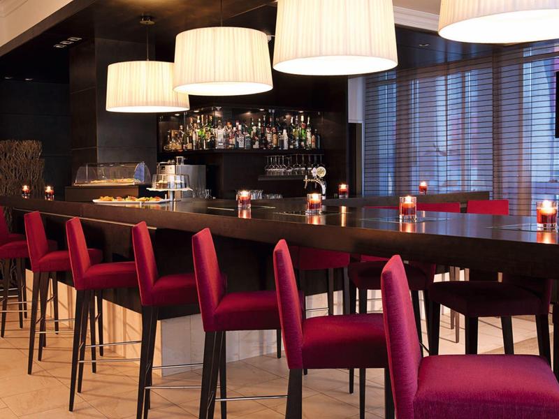 Moderner Barbereich mit langem dunklem Holztheken, roten Barhockern und großen weißen Hängelampen.