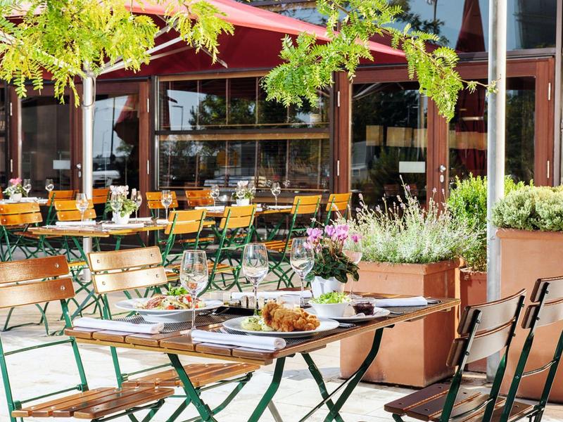 Freiluft-Café mit Holzstühlen, grünen Metallrahmen, Tischen, Pflanzen und Sonnendach.