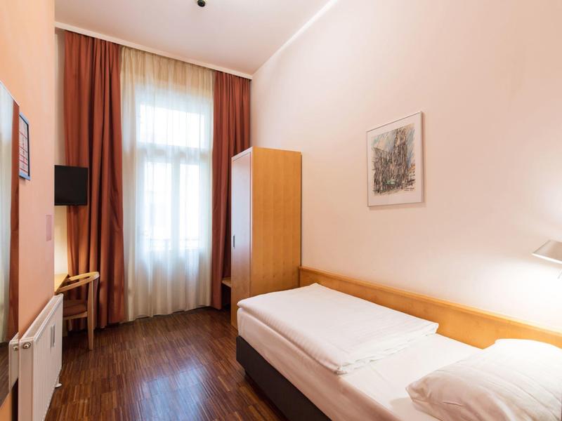 Prosty pokój jednoosobowy z łóżkiem, szafą, krzesłem i oknem z zasłonami.