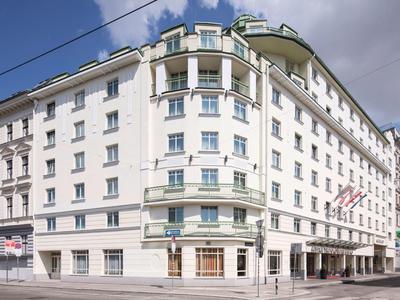 Großes helles Eckgebäude im klassischen Stil mit vielen Fenstern und grünem Dach bei bewölktem Himmel.