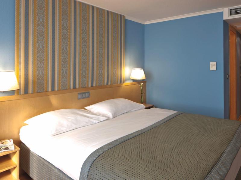 Hotelový pokoj s velkou manželskou postelí, modrou stěnou a dekorativním čelem postele.