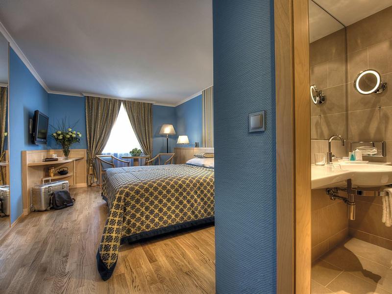 Hotelový pokoj s modrými zdmi, manželskou postelí, televizí a výhledem do koupelny.
