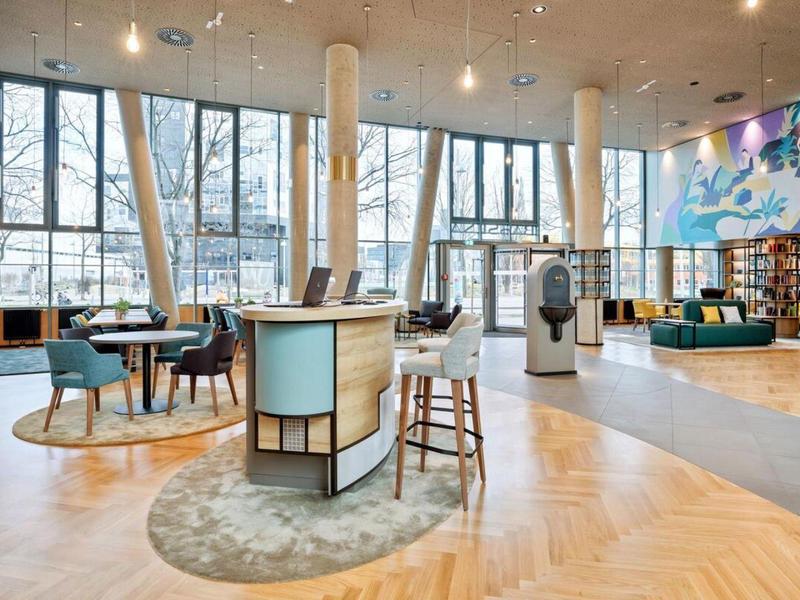 Bright hotel lobby with large windows, seating areas, and a round reception desk.