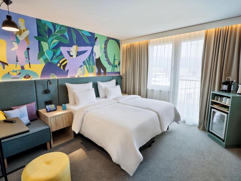 Modern hotel room with two beds, colorful wall art, and balcony with curtains.