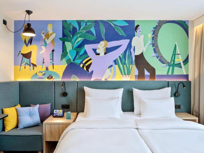 Modern hotel room with two single beds, colorful wall art, and a cozy bench.