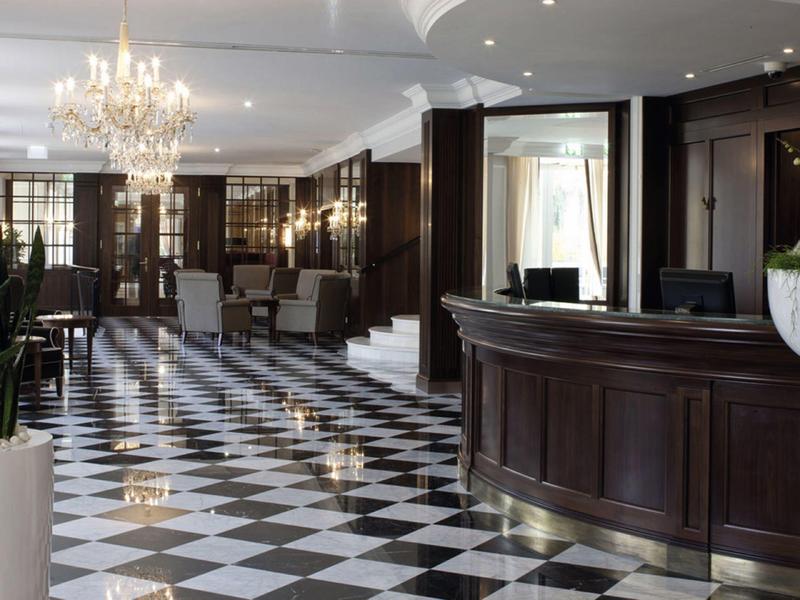 Elegantní hotelová hala s lesklou šachovnicovou podlahou a lustr nad recepcí.