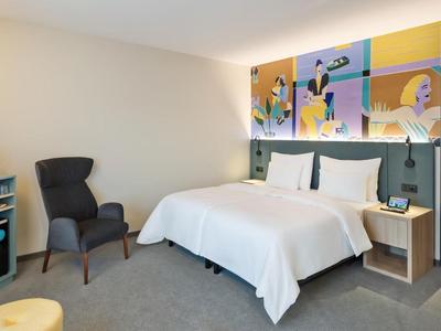 Hotelzimmer mit großem Bett, grauem Sessel, Mini-Kühlschrank und bunter Wandkunst über dem Bett.