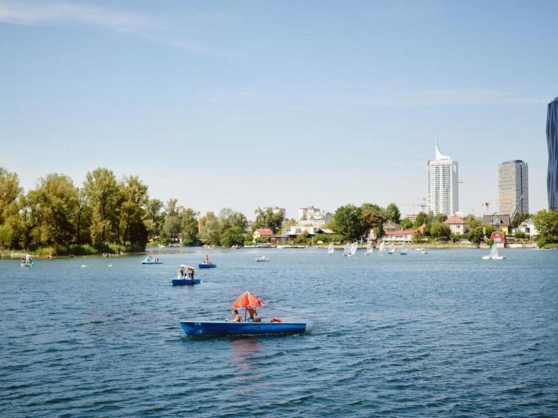 Personas remando en botes azules en un lago con edificios de la ciudad al fondo.