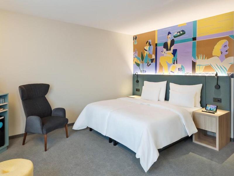 Habitación de hotel moderna con cama doble, sillón cómodo y arte colorido sobre la cabecera.