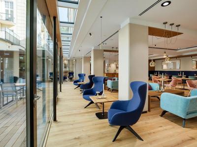 Modern hotel lounge area with blue chairs, wooden floor, and glass front to outdoor space.
