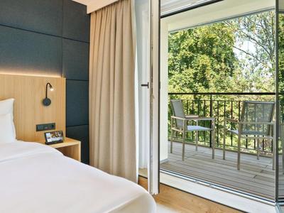 Modern hotel room with bed, nightstand, curtains, and balcony with chairs overlooking trees.