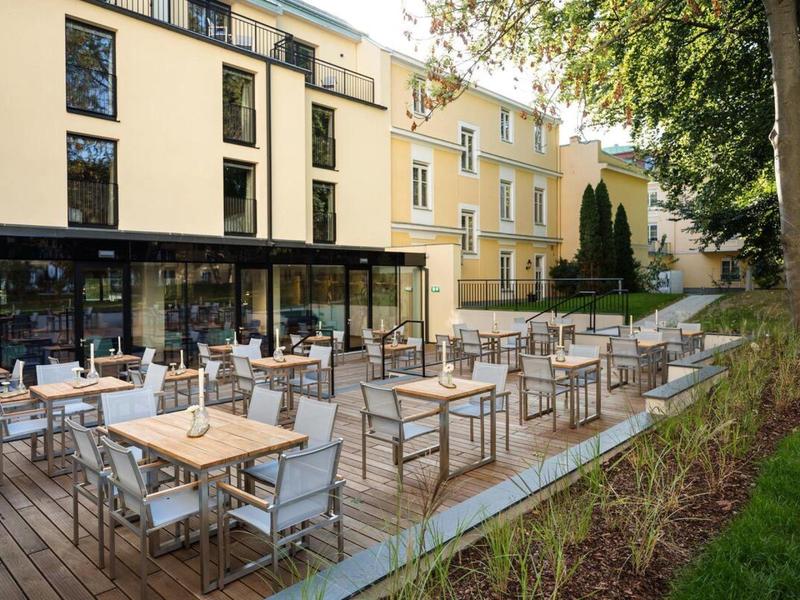 Terasa s stoly a židlemi vedle moderní hotelové budovy a zeleného okolí.