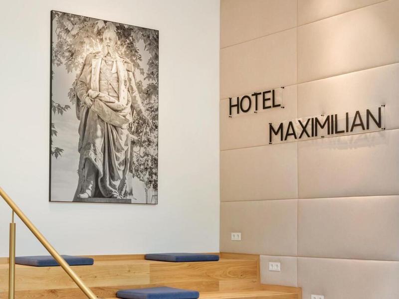 Světlá schodišťová oblast s obrazem na stěně a nápisem 'Hotel Maximilian'.