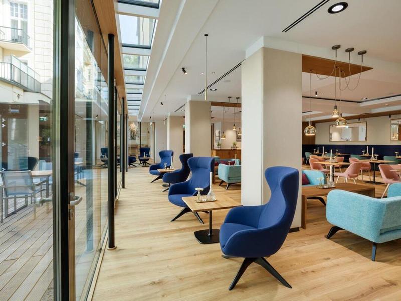 Moderní hotelová lounge s modrým posezením a dřevěnou podlahou vedle velké skleněné fasády.