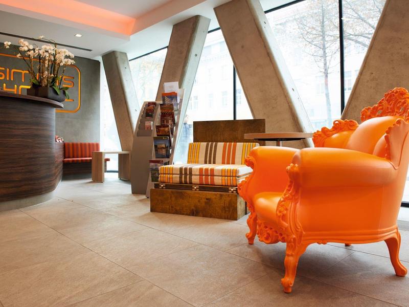 Hall d'hôtel lumineux avec fauteuils baroques orange et meubles en bois élégants près de grandes fenêtres.