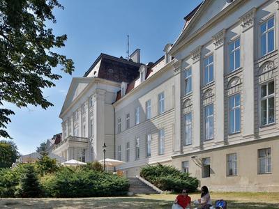 Großes weißes Gebäude mit vielen Fenstern, davor Bäume, Sträucher und mehrere Personen auf einer Wiese.