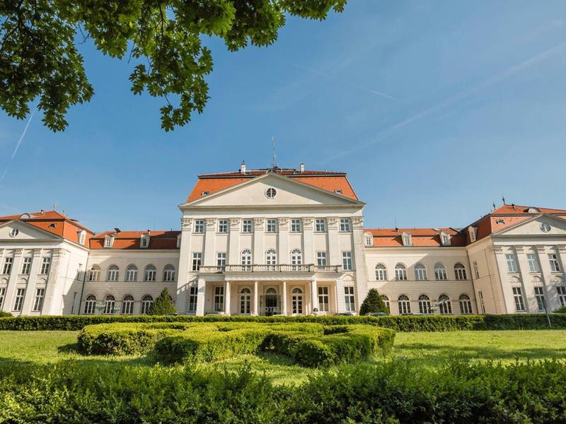 Großes weißes Schloss mit roten Dächern vor blauem Himmel und grünem Garten im Vordergrund.