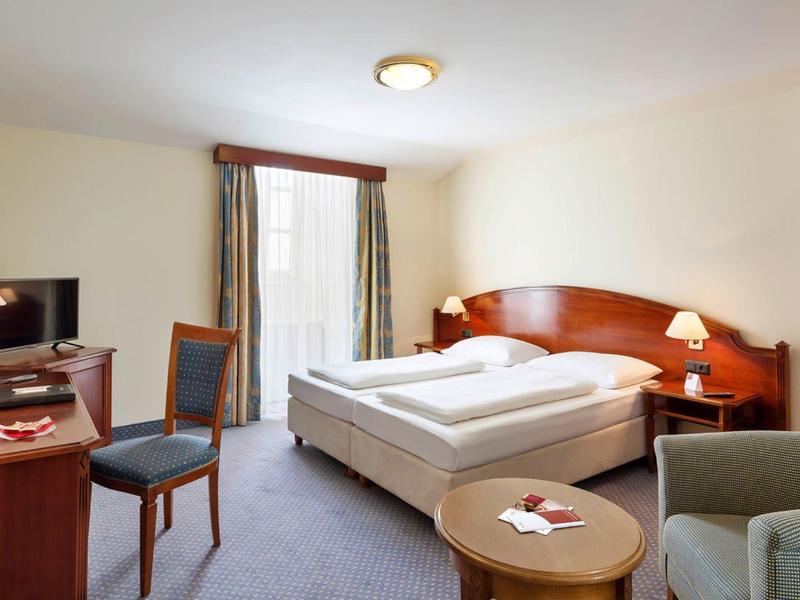 Gemütliches Hotelzimmer mit Doppelbett, Schreibtisch, Stuhl, Sessel und Fenster mit Vorhängen.