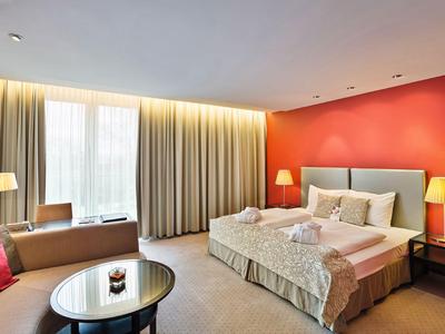 Chambre d'hôtel lumineuse avec lit double, deux tables de chevet et grandes fenêtres avec rideaux.