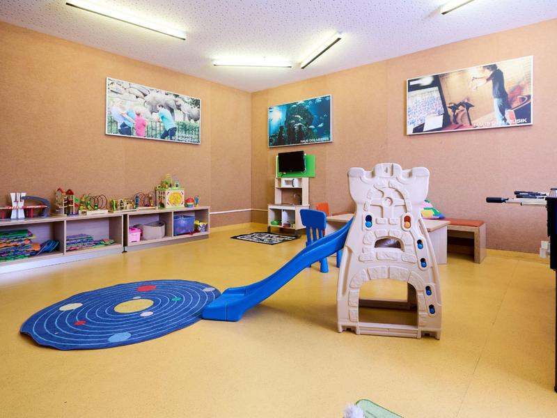 Barevná herna s klouzačkou, naskládanými židlemi a různými hračkami na zdech.