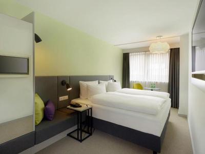 Modern hotel room with double bed, seating area, and large window with curtains.
