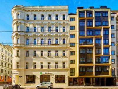 Zwei nebeneinander stehende Gebäude mit hellem Sockel, eines klassisch verziert, das andere modern mit Glas.