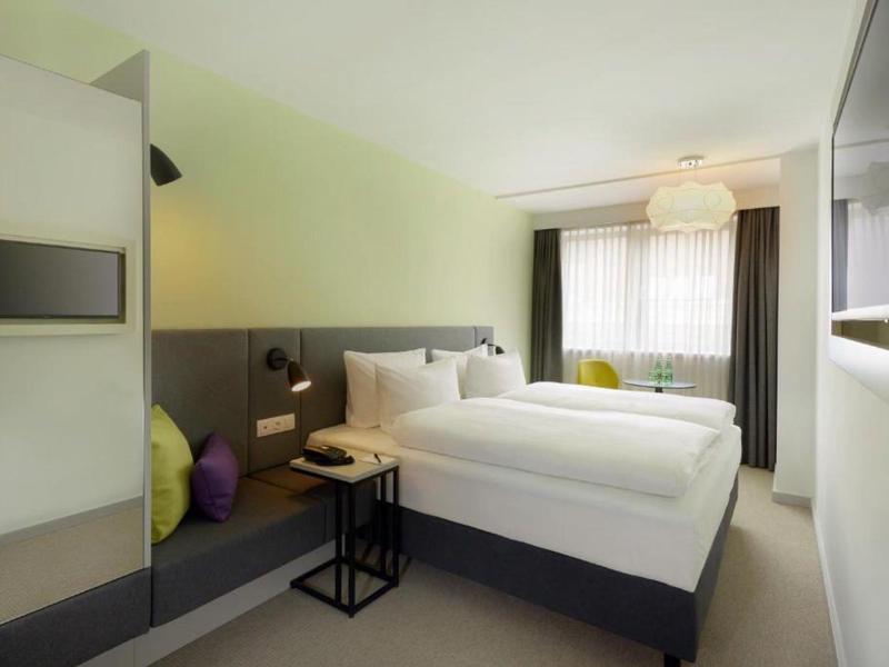 Modern hotel room with double bed, seating area, and large window with curtains.