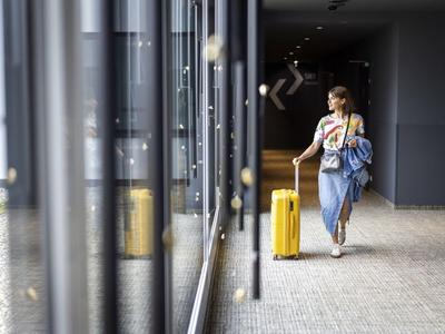 Kobieta z żółtą walizką idzie obok dużych okien w korytarzu hotelowym.