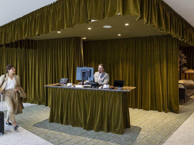 Recepcja hotelu z zielonym zasłoną i gościem z walizką przy ladzie.