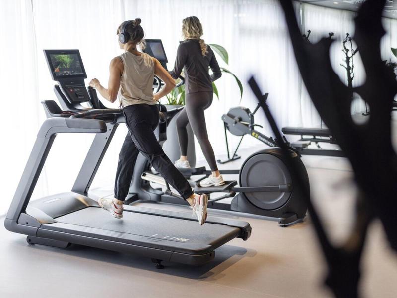 Dwie kobiety biegają na bieżniach w jasnej siłowni z dużymi oknami.