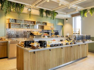 Moderner Frühstücksraum mit langem Buffet, Holzverkleidung und grünen Hängepflanzen am Deckenrand.