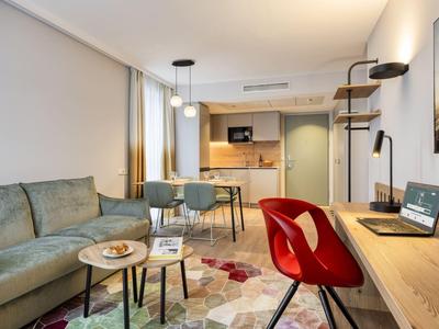 Modern hotel room area with sofa, dining table, red chair, and large desk.