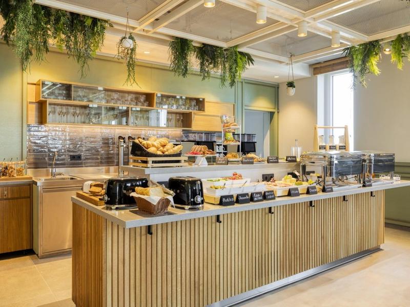 Moderner Frühstücksraum mit langem Buffet, Holzverkleidung und grünen Hängepflanzen am Deckenrand.