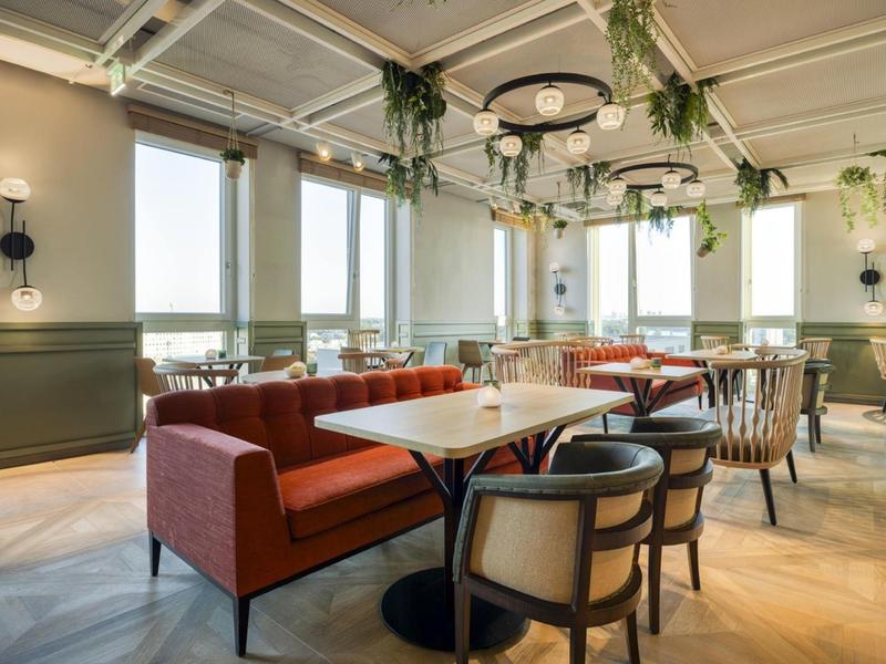 Helles Restaurant mit roten Sitzbänken, Holztischen, Stühlen und großen Fenstern mit Pflanzen.