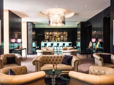 Elegancki hotelowy salon z skórzanymi fotelami i podświetlonym barem w tle.