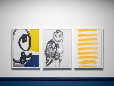 Trois peintures modernes sont accrochées à un mur blanc au-dessus d'une bande bleue.