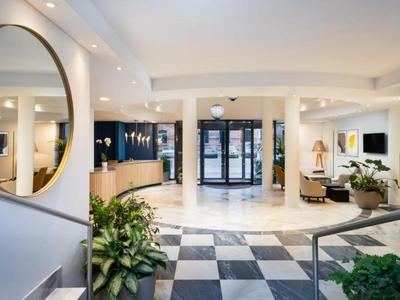 Hall d'hôtel lumineux avec miroir rond, plantes et grandes fenêtres en arrière-plan.